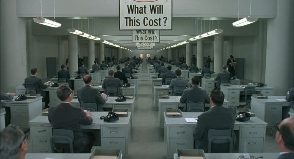 Screenshot from the movie `The Hudsucker Proxy` of an office with lots of people on typewriters and a sign saying `what will this cost?`